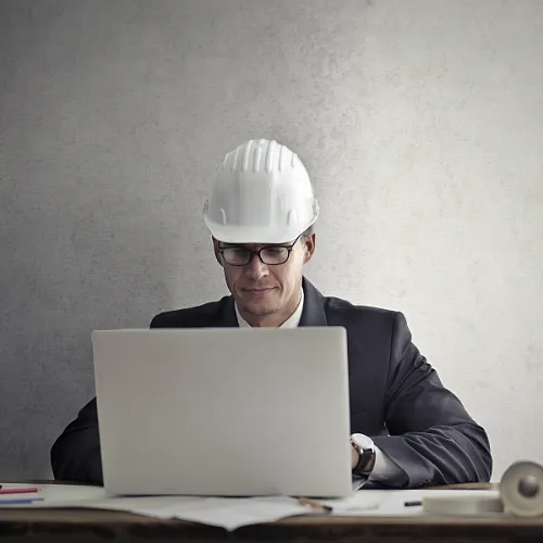 A man in a suit and hard hat is seated at a desk, working on a laptop, showcasing work experience acquired in Canada.