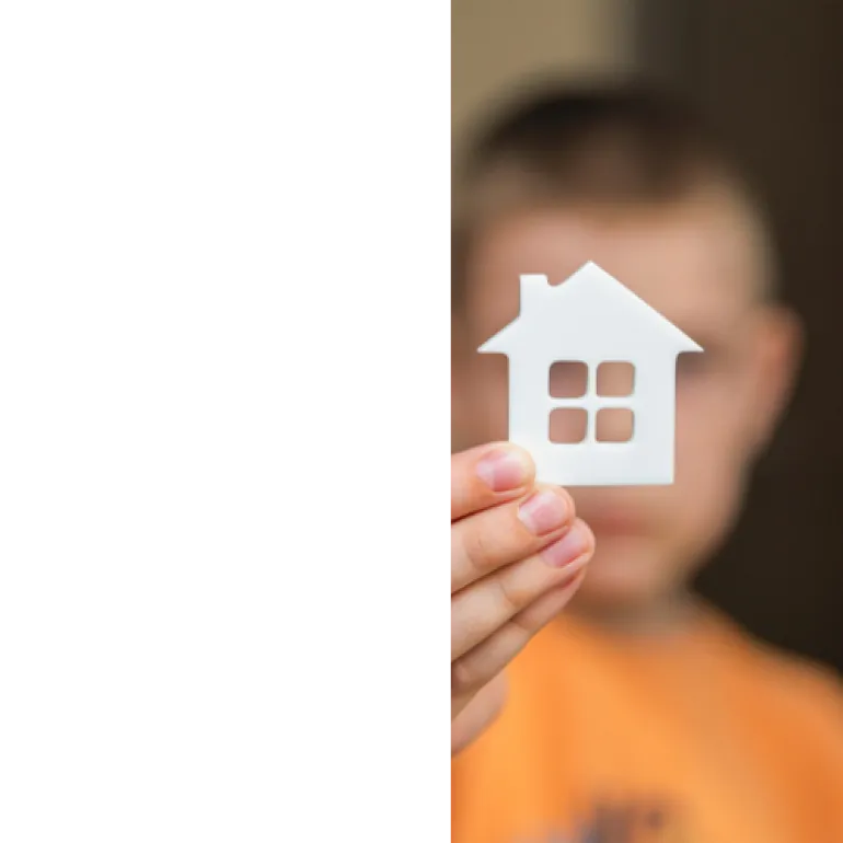 A young child holds a handmade paper house, symbolizing a place to belong.